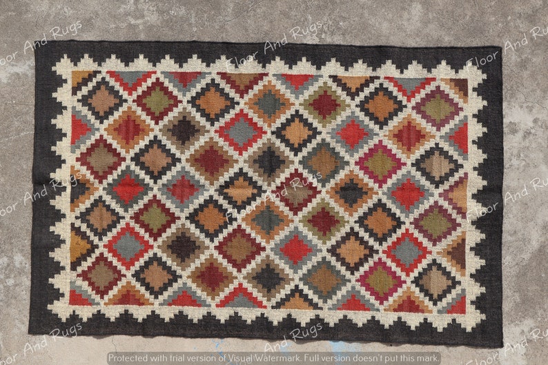 May include: A rectangular woven rug featuring a repeating diamond pattern in shades of red, brown, olive green, gray, and black. The rug is framed by a jagged, off-white border and a solid black outer edge, creating a geometric design.