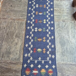 May include: A long, rectangular blue runner rug featuring a white geometric border and a series of small, multi-coloured woven motifs in red, yellow, and green. The rug has a flat-weave texture and is finished with short, light brown fringe at both ends.