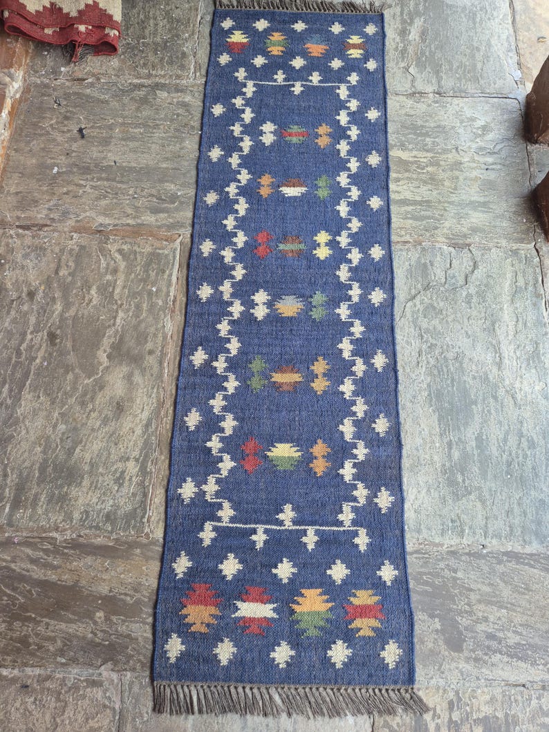 May include: A long, rectangular blue runner rug featuring a white geometric border and a series of small, multi-coloured woven motifs in red, yellow, and green. The rug has a flat-weave texture and is finished with short, light brown fringe at both ends.