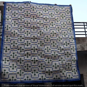 May include: A large rectangular area rug featuring a cream-colored base with a repeating grid pattern outlined in blue. Each grid square contains a central green floral motif with small yellow accents. The rug is finished with a solid blue border.