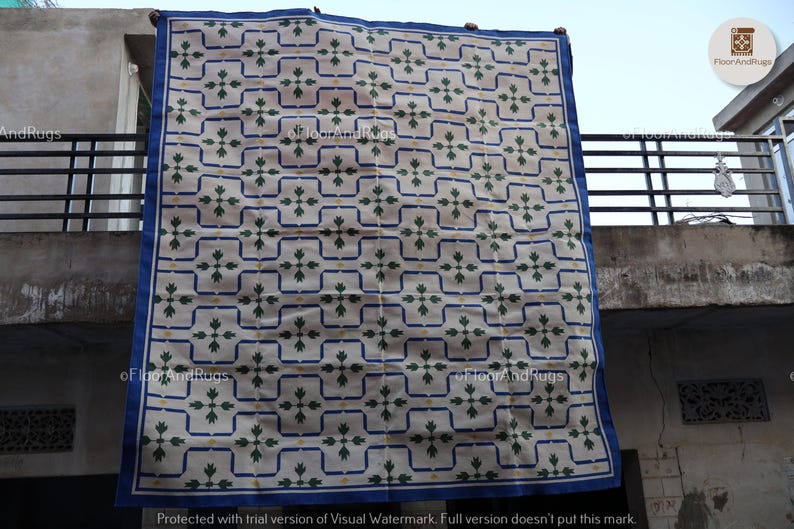 May include: A large rectangular area rug featuring a cream-colored base with a repeating grid pattern outlined in blue. Each grid square contains a central green floral motif with small yellow accents. The rug is finished with a solid blue border.