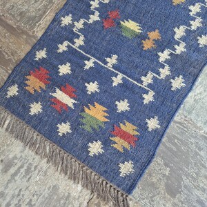 May include: A rectangular blue woven rug featuring a repeating pattern of small white geometric shapes and larger multi-coloured motifs in red, yellow, and green. The rug is finished with a fringe of thin, light brown fibres along the bottom edge.