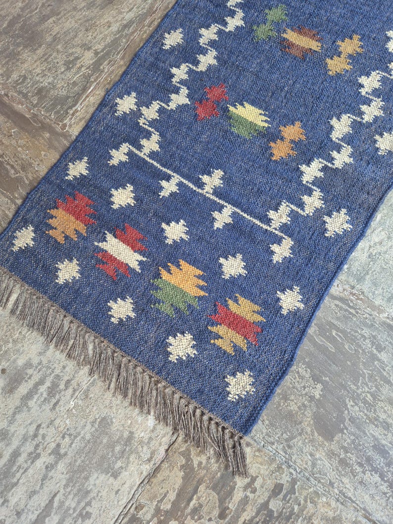 May include: A rectangular blue woven rug featuring a repeating pattern of small white geometric shapes and larger multi-coloured motifs in red, yellow, and green. The rug is finished with a fringe of thin, light brown fibres along the bottom edge.