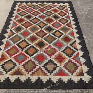 May include: A rectangular area rug featuring a repeating diamond pattern in shades of red, olive green, brown, gray, and black. The rug is bordered by a thick black frame and a white zigzag edge, with the text Floor And Rugs visible on the surface.