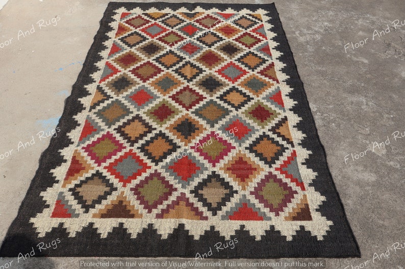 May include: A rectangular area rug featuring a repeating diamond pattern in shades of red, olive green, brown, gray, and black. The rug is bordered by a thick black frame and a white zigzag edge, with the text Floor And Rugs visible on the surface.