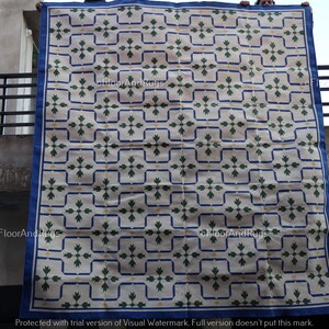 May include: A large rectangular rug featuring a repeating pattern of dark green floral motifs centered within blue outlined squares on a cream background. The rug has a solid blue border and is displayed hanging vertically against a building.