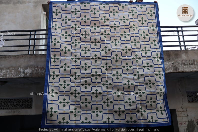 May include: A large rectangular rug featuring a repeating pattern of dark green floral motifs centered within blue outlined squares on a cream background. The rug has a solid blue border and is displayed hanging vertically against a building.