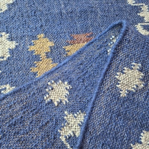 May include: A woven rug featuring a deep blue base colour with a repeating geometric pattern in cream, tan, and red. The textile has a visible coarse texture with a folded corner showing the thickness and the finished edge of the fabric construction.
