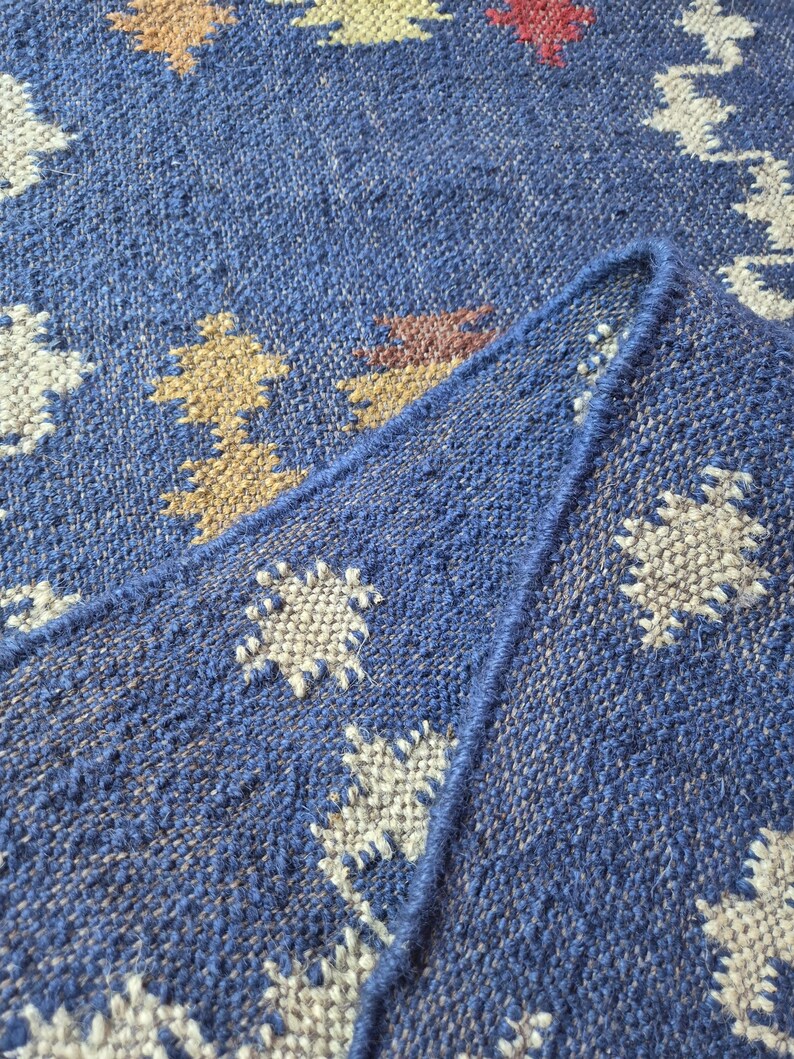 May include: A woven rug featuring a deep blue base colour with a repeating geometric pattern in cream, tan, and red. The textile has a visible coarse texture with a folded corner showing the thickness and the finished edge of the fabric construction.