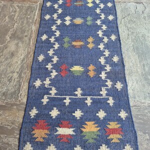 May include: A rectangular blue woven table runner featuring a repeating pattern of cream-coloured geometric shapes and small, multi-coloured motifs in red, yellow, green, and brown. The runner is finished with a fringe along the bottom edge.