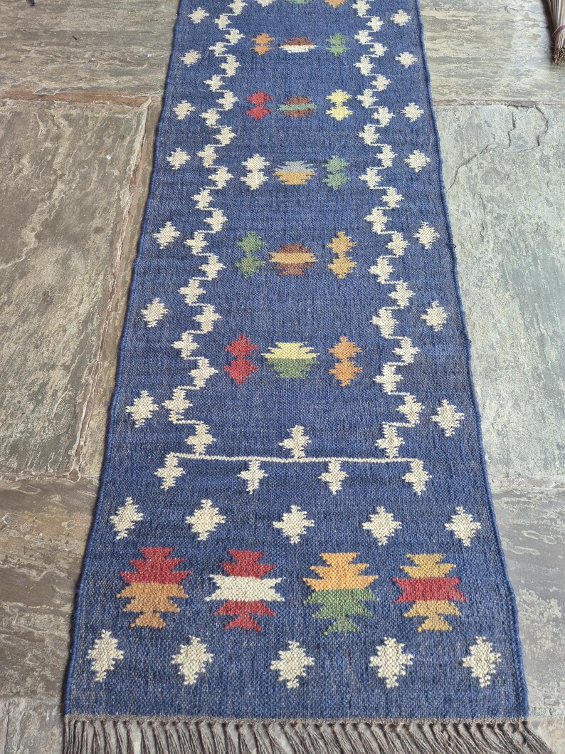 May include: A rectangular blue woven table runner featuring a repeating pattern of cream-coloured geometric shapes and small, multi-coloured motifs in red, yellow, green, and brown. The runner is finished with a fringe along the bottom edge.