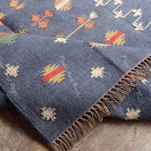 May include: A blue handwoven rug with a geometric pattern in shades of orange, yellow, and white. The rug has fringe on all sides and the text "Floor And Rugs" is visible on the rug.