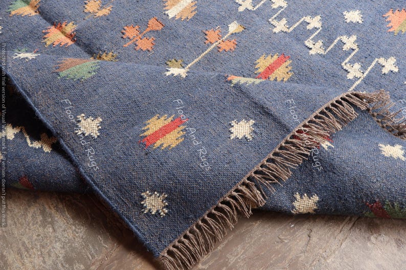May include: A blue handwoven rug with a geometric pattern in shades of orange, yellow, and white. The rug has fringe on all sides and the text "Floor And Rugs" is visible on the rug.