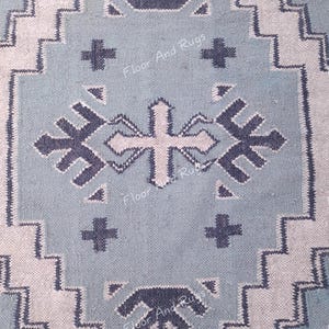 May include: A light blue rug featuring a dark blue geometric pattern with zigzag borders and cross-shaped motifs. The text Floor And Rugs is visible in a repeating pattern across the surface of the woven fabric, which has a textured appearance.