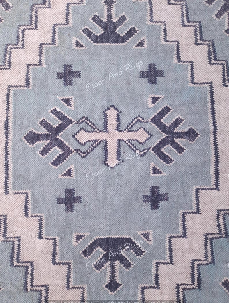 May include: A light blue rug featuring a dark blue geometric pattern with zigzag borders and cross-shaped motifs. The text Floor And Rugs is visible in a repeating pattern across the surface of the woven fabric, which has a textured appearance.