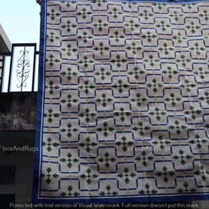 May include: A rectangular area rug featuring a cream-colored base with a repeating grid pattern outlined in blue. Each grid square contains a central green floral motif with four leaves and a small yellow dot, finished with a solid blue border.