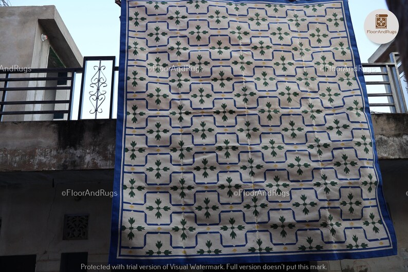 May include: A rectangular area rug featuring a cream-colored base with a repeating grid pattern outlined in blue. Each grid square contains a central green floral motif with four leaves and a small yellow dot, finished with a solid blue border.