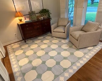 May include: A large area rug featuring a geometric pattern of cream, light blue, and sage green octagons with a matching border. The rug is placed in a room with two striped armchairs, a dark wood dresser, a table lamp, and framed wall art.