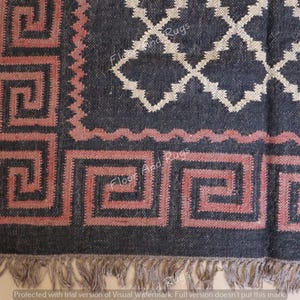 May include: A dark charcoal rug featuring a prominent dusty rose Greek key border pattern and a central white diamond lattice design. The rug is finished with a short, light-colored fringe along the bottom edge, creating a textured appearance.