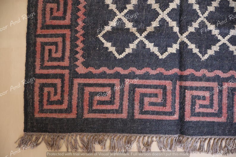 May include: A dark charcoal rug featuring a prominent dusty rose Greek key border pattern and a central white diamond lattice design. The rug is finished with a short, light-colored fringe along the bottom edge, creating a textured appearance.