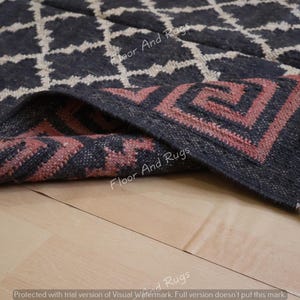May include: A dark charcoal rug featuring a white diamond grid pattern and a folded corner revealing a pink Greek key border design. The rug has a textured surface and short fringe along the edge, with the text Floor And Rugs visible.