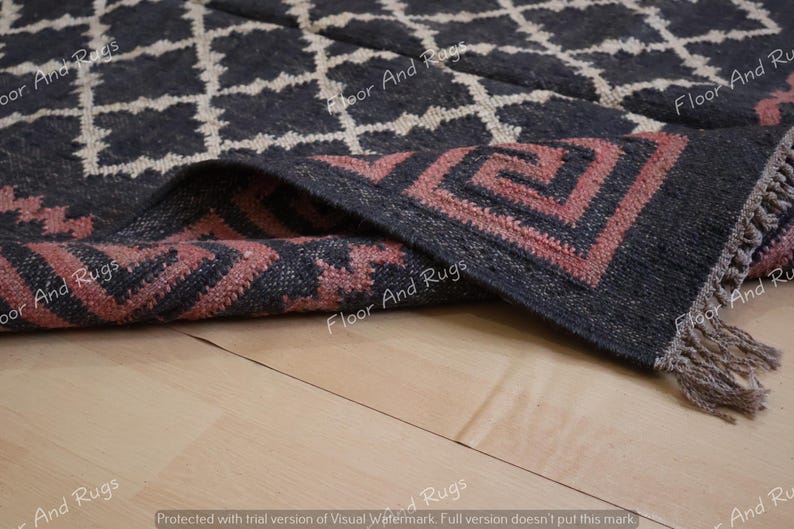 May include: A dark charcoal rug featuring a white diamond grid pattern and a folded corner revealing a pink Greek key border design. The rug has a textured surface and short fringe along the edge, with the text Floor And Rugs visible.