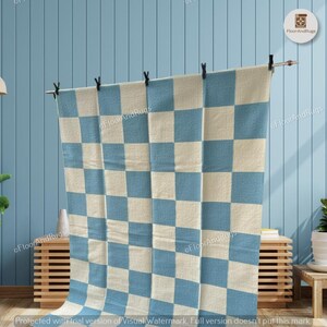 May include: A rectangular rug featuring a repeating checkered pattern of light blue and off-white squares. The rug is suspended from a horizontal metal rod by four black clips, displaying a flat, woven texture across its entire surface area.