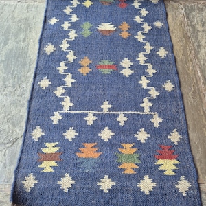 May include: A rolled-up, rectangular blue rug with a geometric pattern in white, orange, yellow, and green. The rug has a fringe on the bottom edge. The design features repeating diamond and stacked shapes.