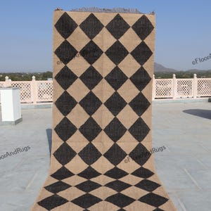 May include: A rectangular area rug featuring a repeating diamond pattern in alternating dark charcoal and natural tan colours. The rug has a textured, woven appearance with short, frayed fringe detailing along the top and bottom edges.