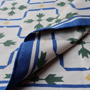May include: A woven rug featuring a white base with a repeating geometric pattern in blue and green. The design includes blue outlined squares, green leaf-like motifs, and small yellow accents. The rug is folded over to reveal the underside texture.