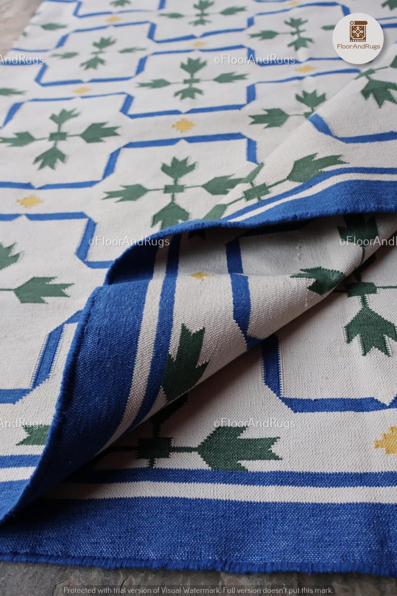 May include: A woven rug featuring a white base with a repeating geometric pattern in blue and green. The design includes blue outlined squares, green leaf-like motifs, and small yellow accents. The rug is folded over to reveal the underside texture.