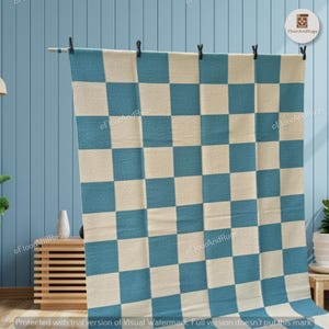 May include: A rectangular area rug featuring a repeating checkered pattern of alternating teal blue and off-white squares. The rug is suspended from a horizontal rod by five black clips, displaying a flat, woven texture across its entire surface.