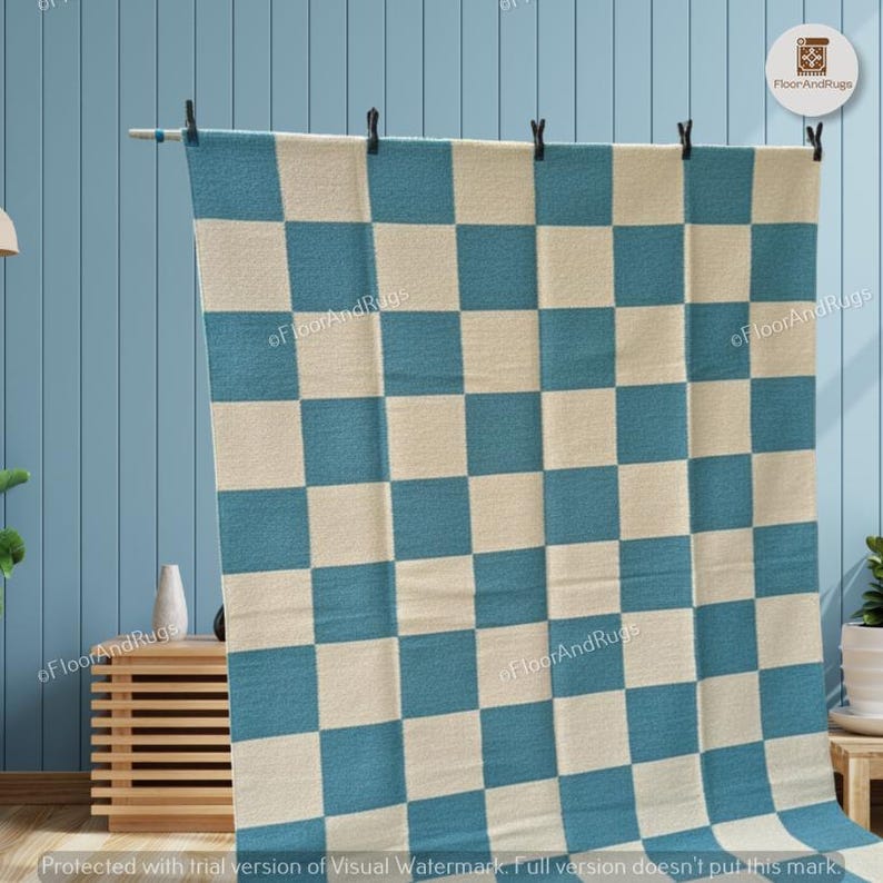 May include: A rectangular area rug featuring a repeating checkered pattern of alternating teal blue and off-white squares. The rug is suspended from a horizontal rod by five black clips, displaying a flat, woven texture across its entire surface.