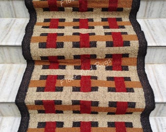 May include: A rectangular woven runner rug featuring a geometric grid pattern in red, tan, brown, and black. The rug has a dark brown border and fringe at the ends. The text Floor And Rugs is visible in white across the surface of the woven runner.