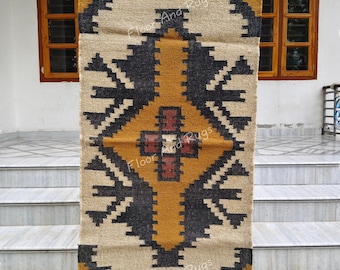May include: A rectangular woven rug featuring a geometric pattern in charcoal, mustard yellow, and cream. The design includes a central diamond motif with a dark red cross element. The rug has fringed edges and the text Floor And Rugs is visible.