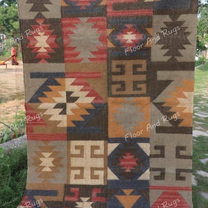 Puede incluir: Una alfombra rectangular con un patrón geométrico en tonos marrón, rojo, beige y azul. La alfombra presenta un diseño de patchwork con varias formas y motivos, incluyendo diamantes y cruces. La alfombra se exhibe al aire libre sobre una superficie cubierta de hierba.