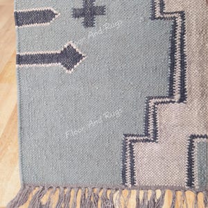 May include: A woven rug in light blue with dark gray geometric patterns, including arrow shapes and a cross. The rug features a stepped border design in light gray and dark gray, finished with a fringe of light gray tassels along the bottom edge.