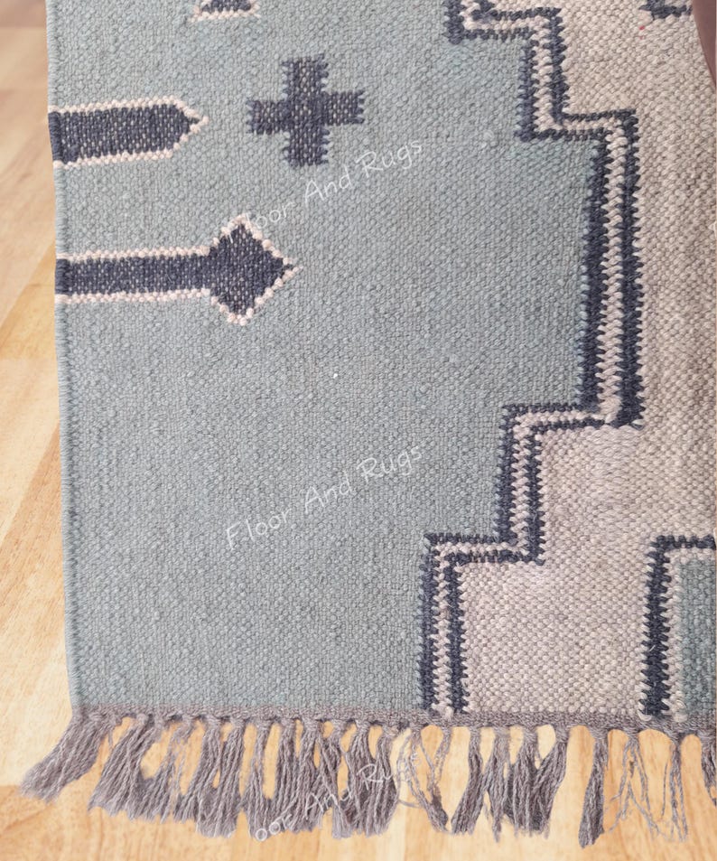 May include: A woven rug in light blue with dark gray geometric patterns, including arrow shapes and a cross. The rug features a stepped border design in light gray and dark gray, finished with a fringe of light gray tassels along the bottom edge.