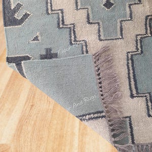 May include: A woven area rug featuring a geometric pattern in shades of light blue, cream, and dark charcoal gray. The rug has a folded corner revealing a solid light blue underside and a fringe edge. The text Floor And Rugs is visible.