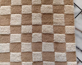 May include: A rectangular rug featuring a repeating checkered pattern of alternating light beige and tan squares. The surface has a thick, tufted, and textured pile construction, creating a distinct grid design across the entire fabric area.