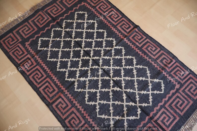 May include: A rectangular area rug featuring a dark charcoal background with a central white diamond lattice pattern. The border displays a repeating pink Greek key motif. The rug has short fringe on the top and bottom edges.