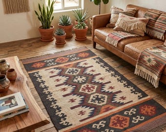 May include: A rectangular area rug featuring a geometric pattern in shades of cream, dark brown, rust orange, and muted blue. The rug has a central diamond motif, bordered by repeating hexagonal shapes and finished with fringe on both short ends.