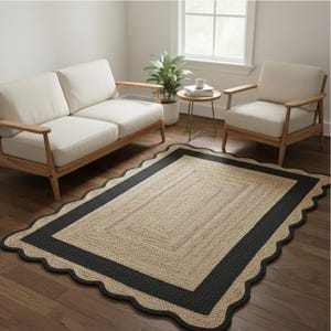 May include: A rectangular jute rug with a scalloped black border and a natural beige center. The rug has a woven texture and is placed on a hardwood floor. The room also features a white sofa, chair, and side table.