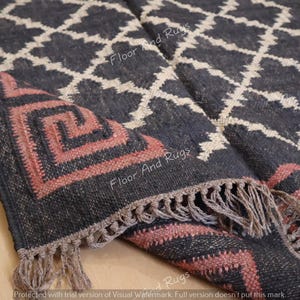 May include: A woven area rug featuring a dark charcoal background with a cream-colored diamond lattice pattern. The border displays a salmon-colored Greek key motif. The edges of the rug are finished with a short, knotted fringe in a neutral tone.