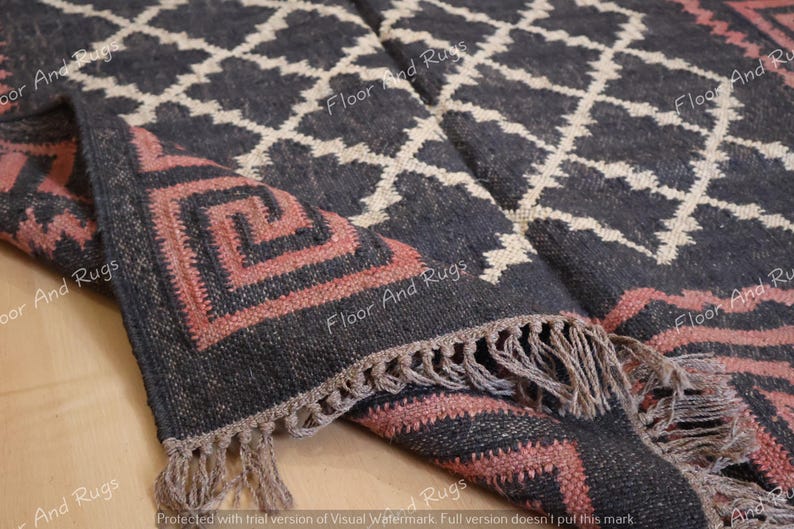 May include: A woven area rug featuring a dark charcoal background with a cream-colored diamond lattice pattern. The border displays a salmon-colored Greek key motif. The edges of the rug are finished with a short, knotted fringe in a neutral tone.