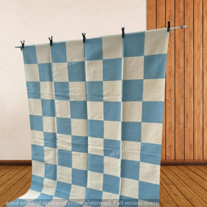 May include: A rectangular rug featuring a repeating checkered pattern in alternating light blue and off-white squares. The textile is suspended from a horizontal rod by four black clips, displaying a flat, woven texture throughout the entire surface.
