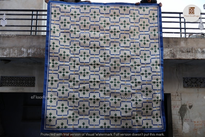 May include: A large rectangular area rug featuring a cream-colored background with a repeating grid pattern of dark blue lines. Each grid square contains a centered green floral motif with small yellow accents. The rug has a solid dark blue border.