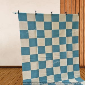 May include: A rectangular rug featuring a repeating checkerboard pattern in alternating teal blue and off-white squares. The textile is suspended from a horizontal rod by four black clips, displaying a flat, woven texture throughout the surface.
