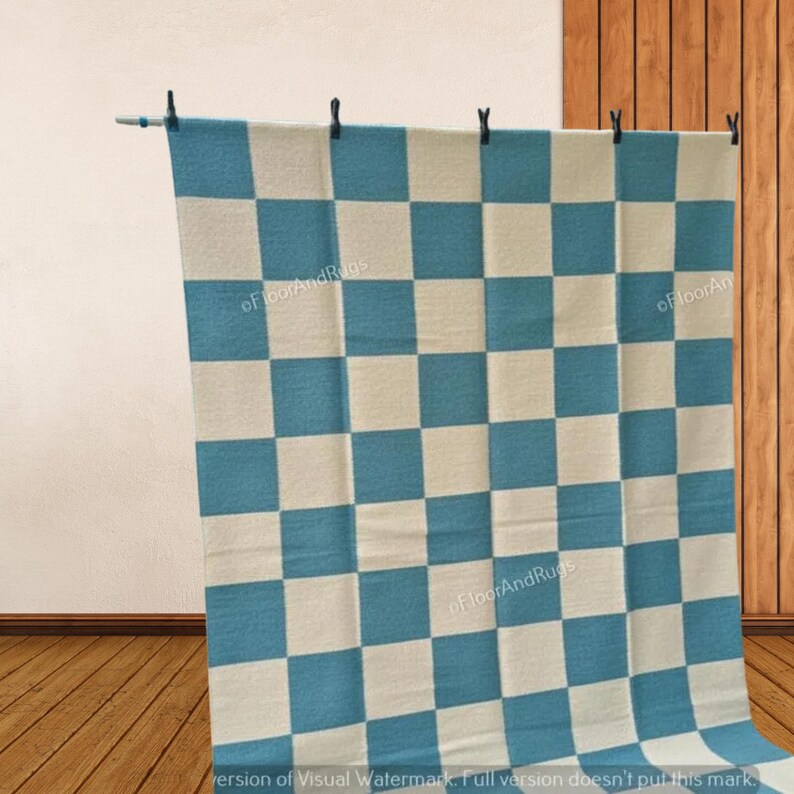 May include: A rectangular rug featuring a repeating checkerboard pattern in alternating teal blue and off-white squares. The textile is suspended from a horizontal rod by four black clips, displaying a flat, woven texture throughout the surface.