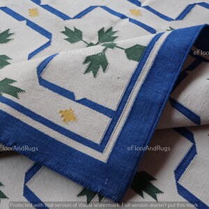 May include: A woven area rug featuring an off-white base with a repeating geometric pattern in blue, dark green, and yellow. The design includes blue outlined octagonal shapes, green leaf-like motifs, and small yellow cross-shaped accents.